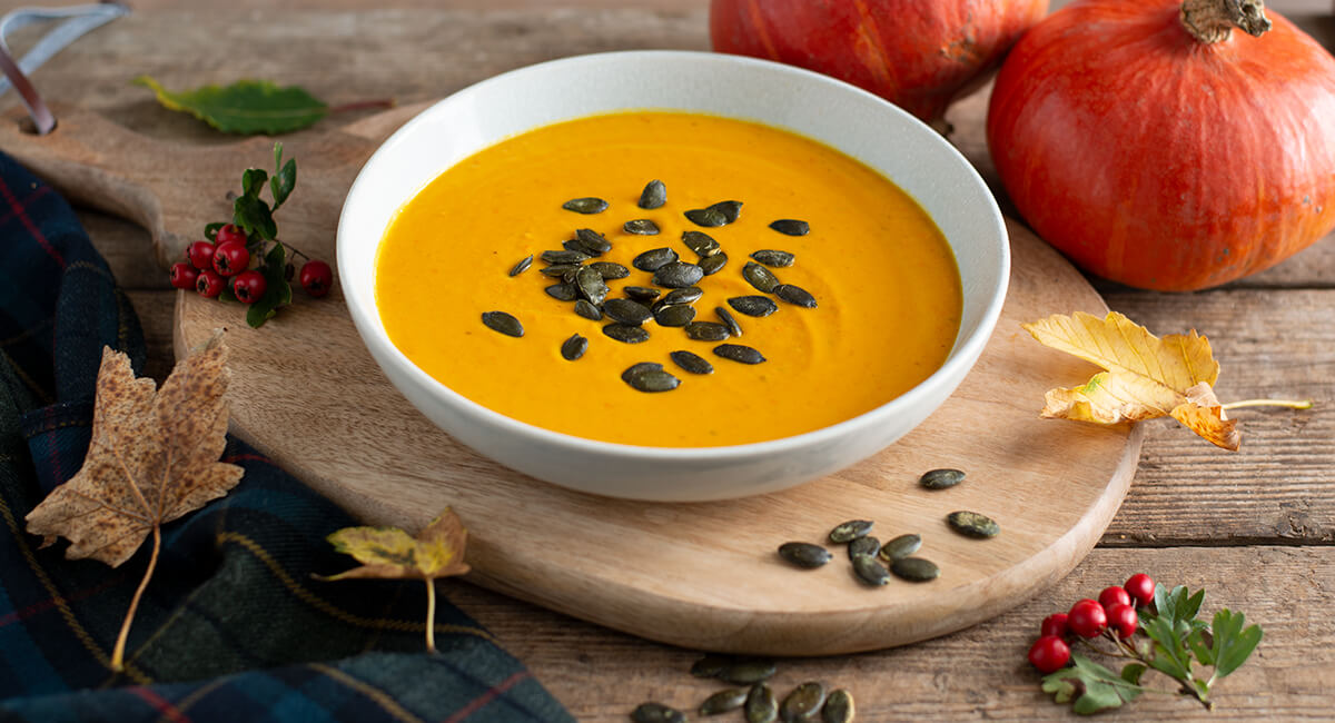 Dýňová polévka na talíři, dýňová semínka, dřevěný stůl a kolem dýně a podzimní listy